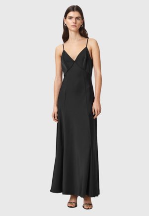 Femme debout portant une longue robe noire sans manches avec de fines bretelles et des sandales noires à bout ouvert sur un fond neutre.