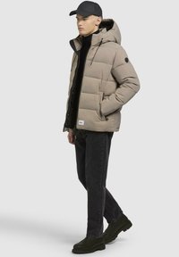 Polstret beige jakke med hætte, sorte accenter, sidelommer, lynlås foran, båret over en sort turtleneck og mørke jeans, med grøn fodtøj.