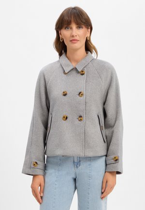 Frau trägt eine graue, doppelreihige Wolljacke mit braunen Knöpfen und hellblauen Jeans, steht vor einem schlichten weißen Hintergrund.