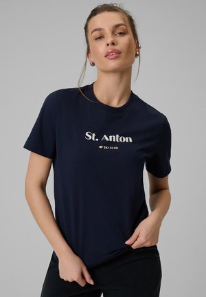 Młoda kobieta w granatowej koszulce "St. Anton Ski Club", trzymająca dolną krawędź i patrząca lekko do góry na szarym tle.