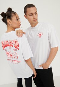 Hvide t-shirts med grafiske tryk i r�ød. Den ene skjorte viser "SHAKE IT UP" og tegnefilmsfigurer, mens den anden har et mindre grafisk tryk på brystet.