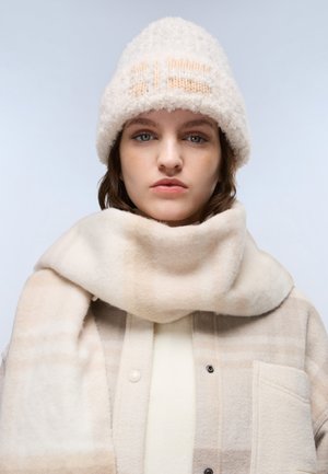 Weiße, flauschige Strickmütze mit Pfirsichakzenten, geschichteter, weicher beiger Schal und kariertes, beiges Oberteil mit großen aufgenähten Taschen.