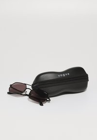 Óculos de sol pretos com lentes escuras repousando ao lado de uma caixa rígida preta com fecho de zip e o logotipo "vogue" em branco.