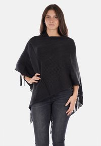Poncho nero lavorato a maglia con un motivo testurizzato, caratterizzato da orli frangiati e un design loose e asimmetrico. Indossato su jeans scuri.