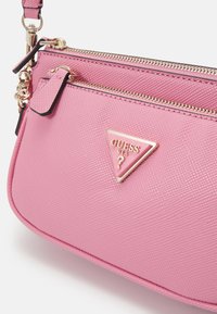 Růžový texturovaný kožený náramek Guess se zlatými zipy a trojúhelníkovou logovou destičkou na přední kapse.