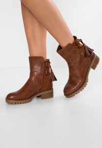 Bottines marron en cuir synthétique, avec une fermeture éclair sur le côté, des accents de franges, un talon épais et une semelle en caoutchouc texturé.