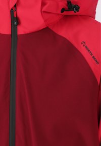 Rød og bordeaux jakke med fuld lynlås, der har høj krave, justerbar hætte og sort logoaccent. Glat tekstur og letvægtsdesign.