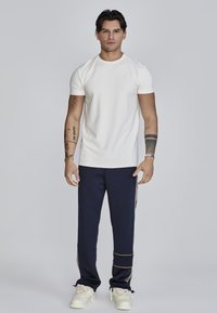 Camiseta blanca de manga corta con un logo sutil, combinada con pantalones deportivos azul marino con rayas doradas y puños elásticos. El calzado son zapatillas color crema.