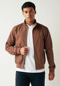 Chaqueta bomber marrón con cierre de cremallera en el frente, ajuste casual, puños y dobladillo acanalados, con un pequeño logotipo bordado en el pecho. Lleva una camiseta blanca debajo.
