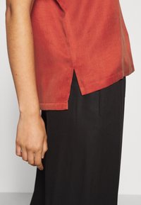 Rustrød skjorte med struktureret overflade og sideslids, stylet med sorte bukser. Skjorten har en afrundet kant og diskrete sømme.