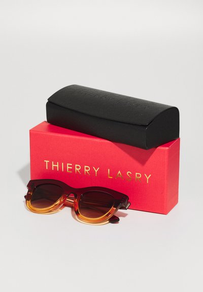 Lunettes de soleil avec un design en écaille de tortue bicolore, présentant des montures supérieures marron foncé et des montures inférieures ambre clair, à côté d'une boîte rouge et noire.