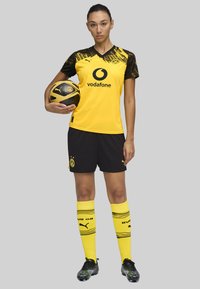 Divisa calcistica del Borussia Dortmund con una maglia gialla con dettagli neri, pantaloni corti neri e calze gialle con motivo a righe. Include un pallone da calcio.