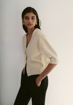 Vrouw met donker haar, draagt gouden oorringen in de vorm van ringen, een crème V-hals cardigan met korte mouwen en een zwarte broek, staand tegen een witte muur.