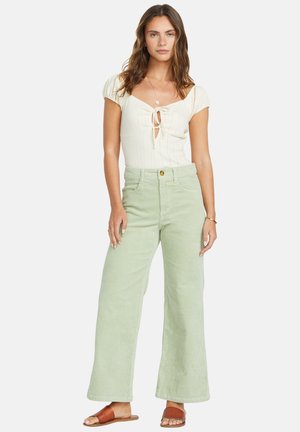 Donna che indossa un top corto a maniche corte color crema con nodo frontale, pantaloni in corduroy larghi di colore verde chiaro e sandali scivolati marroni, in piedi contro uno sfondo bianco.