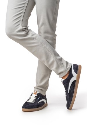 Baskets en daim bleu marine avec des accents blancs, des panneaux texturés et des semelles en caoutchouc beige, associées à un pantalon ajusté gris clair.