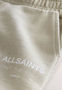 Tessuto beige con "ALLSAINTS" stampato in bianco, accompagnato da caratteri giapponesi più piccoli e dalla parola "LONDON" sotto.