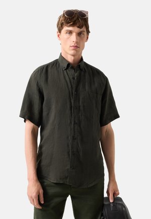 Junger Mann mit schwarzem kurzärmligem Hemd mit Knöpfen und Sonnenbrille auf dem Kopf, hält eine schwarze Ledertasche vor einfarbigem Hintergrund.