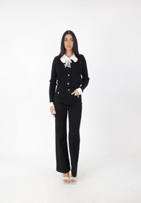 Cárdigan negro de punto con cuello blanco y detalle de lazo, combinado con pantalones vaqueros negros acampanados y sandalias de tiras de color claro.