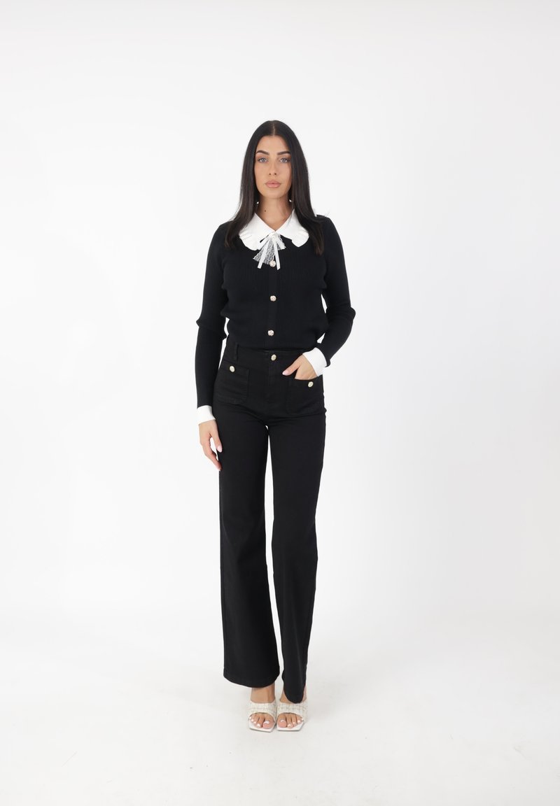 Cárdigan negro de punto con cuello blanco y detalle de lazo, combinado con pantalones vaqueros negros acampanados y sandalias de tiras de color claro.