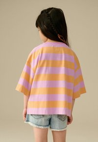 T-shirt oversized à manches courtes avec des rayures alternées orange et lavande, associé à un short en denim effiloché. Vue arrière montrant des cheveux tressés.