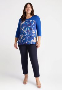 Niebieska bluzka o długości trzech czwarte z kwiatowym wzorem w odcieniach niebieskiego na jasnym tle, zestawiona z ciemnonawęglowymi spodniami slim-fit.
