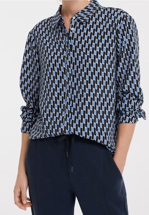 Chemise à manches longues boutonnée avec un motif géométrique bleu et noir, associée à un pantalon bleu foncé, une main dans la poche, sur fond blanc.