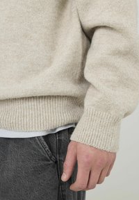 Pull beige en maille avec ourlet et poignets côtelés, fabriqué dans un tissu doux et texturé ; porté sur une chemise blanche et accompagné d'un jean gris.
