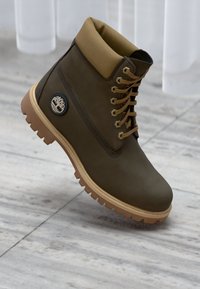 Cizme impermeabile de culoare verde închis, cu un guler din țesătură bej, urechi metalice și o talpă din cauciuc texturată. Prezintă o etichetă logo pe partea laterală.
