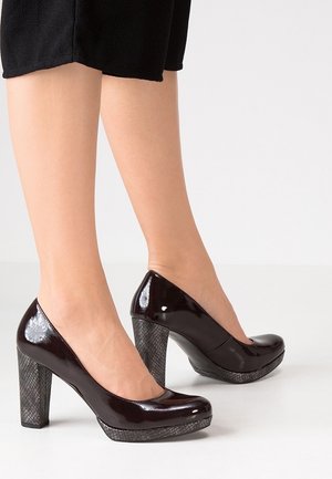 Femme portant des chaussures à talons hauts brillantes brun foncé avec une plateforme texturée et un talon bloc, debout sur un sol blanc, jupe noire visible.