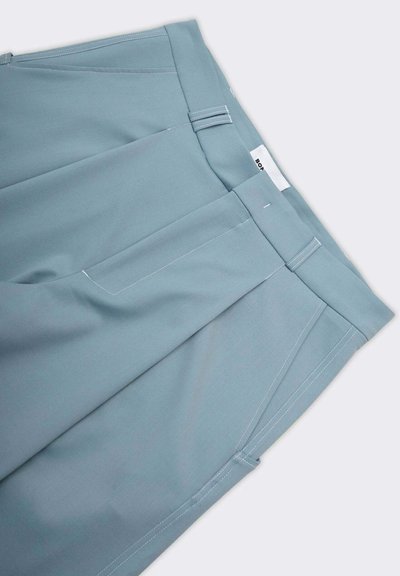 Pantaloni azzurri chiari con una texture liscia, pieghe frontali, tasche laterali e dettagli di cucitura bianca. Vita piatta con etichetta visibile.