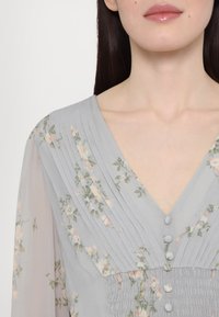 Blouse gris clair en tissu transparent, avec motifs floraux, design plissé, encolure en V et boutons décoratifs à l'avant.