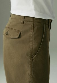 Shorts en coton vert olive avec une poche latérale et une poche arrière à bouton, présentant un tissu texturé et des détails cousus.
