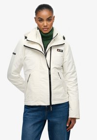 Biała, ocieplana kurtka z zamkiem błyskawicznym z przodu i regulowanym kapturem. Posiada czarne zamki, kieszenie oraz naszywkę z logo na klatce piersiowej.