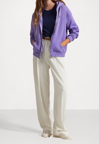 Felpa zip-up viola, pantaloni bianchi a gamba larga e sneaker bianchi. Presenta tasche e una vestibilità rilassata con un piccolo logo sulla felpa.