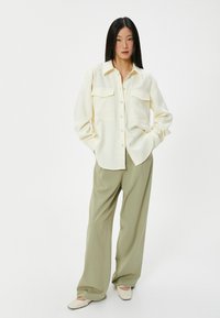 Camicia button-up crema chiaro con due tasche sul petto, abbinata a pantaloni larghi verde oliva e scarpe beige slip-on.