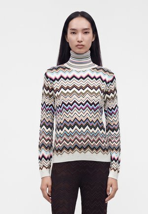 Donna con capelli lunghi neri che indossa un maglione a collo alto con motivi a zigzag multicolore e pantaloni scuri con motivo a zigzag su sfondo bianco.