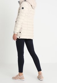 Beige pufferjakke med hætte, lodret syninger og logodetalje. Båret med mørke slim fit jeans og lyse sneakers. Glat tekstur.