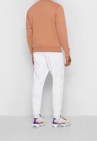 Ljust brunt sweatshirt med ribbad mudd och nertill, i kombination med vita joggingbyxor med dekorativ sidopanel och sneakers med lila detaljer.