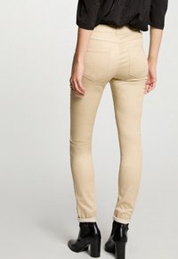 Beige slim-fit broek met een middelhoge taille, voorzien van achterzakken en opgerolde manchetten. Gecombineerd met zwarte enkellaarsjes. Soepele textuur.