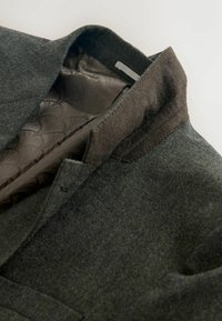 Chaqueta de lana verde oscuro con un acabado texturizado, forro interior suave y con patrón, y un cuello estructurado.