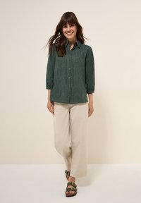 Chemise boutonnée vert foncé à manches ¾, avec un motif texturé. Associée à un pantalon large beige clair et des sandales marron.