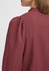 Bordeaux-farbenes, strukturiertes Oberteil mit gefalteten Schultern und einem Rüschenausschnitt. Besteht aus weichem, zerknittertem Stoff mit dezenten horizontalen Linien.