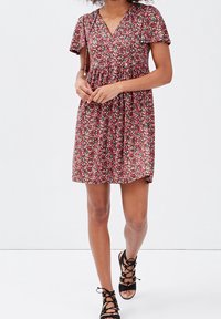 Femme marchant dans une robe courte à fleurs rouges avec un col noué à volants et des manches courtes, associée à des sandales noires à lacets.