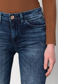 Slim-fit tmavě modré džíny vyrobené z denimu. Obsahují klasické kapsy, měděné zapínání na knoflík a jemné blednutí podél stehen.