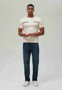 Homem em pé, vestindo uma t-shirt branca com riscas verdes, jeans azuis e ténis brancos, contra um fundo simples.