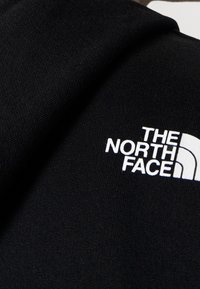 Matériau en polaire noire avec un logo brodé blanc. Le design comprend un accent graphique incurvé et une surface de tissu texturée.