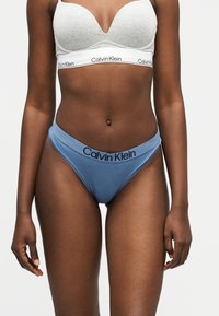 Γυναίκα που φοράει ανοιχτό γκρι σουτιέν Calvin Klein και μπλε εσώρουχα Calvin Klein, στέκεται με τα χέρια χαλαρά στα πλευρά μπροστά σε απλό φόντο.