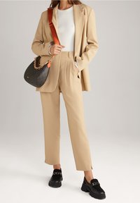Beżowa marynarka zapinana na guziki i pasujące do niej spodnie, zestawione z białym prążkowym topem i czarnymi masywnymi loafersami. W komplecie znajduje się brązowa i czarna torba na ramię.