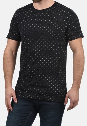 Homme portant un t-shirt noir à manches courtes avec de petits motifs blancs et un jean bleu foncé, debout les mains détendues le long du corps.