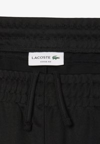 Zwarte ruime broeken met een elastische tailleband en koord. Voorzien van een label met "LACOSTE" en een groene krokodillogo. Gladde textuur.